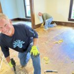 Two men are removing old flooring in an empty room, wearing gloves and using tools. One is smiling at the camera while kneeling on the floor.