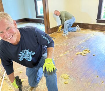 Two men are removing old flooring in an empty room, wearing gloves and using tools. One is smiling at the camera while kneeling on the floor.