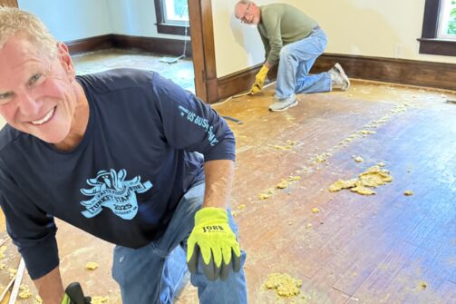 Two men are removing old flooring in an empty room, wearing gloves and using tools. One is smiling at the camera while kneeling on the floor.