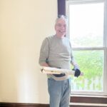 Smiling man in sunglasses and gloves stands by a window holding window blinds, wearing a gray shirt and jeans.