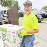Man wearing a neon yellow shirt and gloves, smiling while holding paper boxes outdoors near a truck on a residential street.