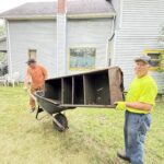 Two men moving a rusty metal shelf in a wheelbarrow outside near a gray house with yellow window trim.