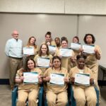A group of women in tan uniforms proudly holding certificates, posing with a man in casual clothes in a bright indoor room.