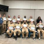 A group of people in tan uniforms proudly holding certificates, with two others standing beside them in a classroom setting. Everyone’s smiling for the camera.