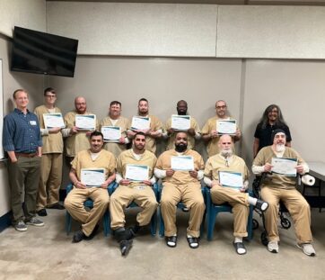 A group of people in tan uniforms proudly holding certificates, with two others standing beside them in a classroom setting. Everyone’s smiling for the camera.