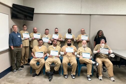 A group of people in tan uniforms proudly holding certificates, with two others standing beside them in a classroom setting. Everyone’s smiling for the camera.