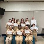 A group of women in tan uniforms proudly holding certificates, posing with a man in a white shirt in a classroom-like setting.