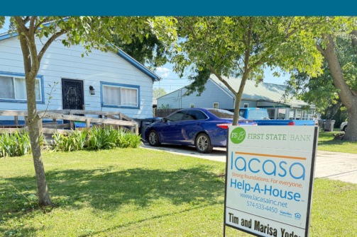 Blue house with a ramp, a car in the driveway, and a yard sign for Help-A-House, featuring a quote about feeling safe at home.
