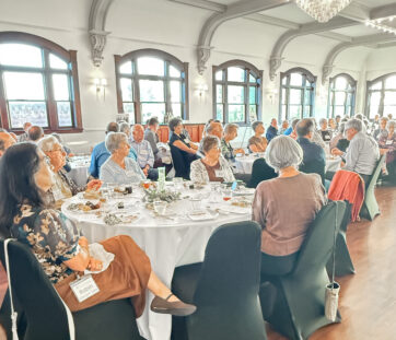 A group of older adults sit around tables at a banquet in a bright, elegant room, listening to a speaker.