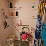 Shower stall with a shower chair, pink towel, hygiene bottles, wind chime, and cleaning supplies. Blue floral shower curtain and grab bar are visible.