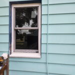 A white-trimmed window on a light blue house with tools nearby. The reflection of trees is visible in the glass.