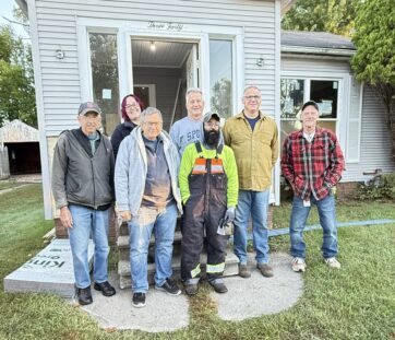 Seven people smiling and standing in front of a small house on a sunny day, looking ready for some work or a group project.