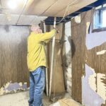 Man in yellow shirt and mask working on basement renovation, removing damaged ceiling panels. Old paneling and debris scattered on floor, room has exposed pipes and wood walls.