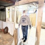 A person wearing a mask and gloves carries boards in a messy basement under renovation, dressed casually in jeans and a sports sweatshirt.