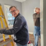 Two men are working on a home improvement project, one standing near a yellow ladder and smiling, the other focused on the wall.