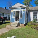 A small house with blue siding is undergoing renovations. A black pickup truck is parked nearby and trimmed bushes are piled on the grass.