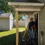 A man in work gloves stands by a porch, looking up, surrounded by tools and wooden planks, on a sunny day next to a house and garage.