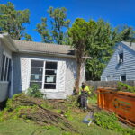 A worker in a yellow vest is clearing branches in front of a house, next to an orange dumpster, with clear blue sky and trees in the background.